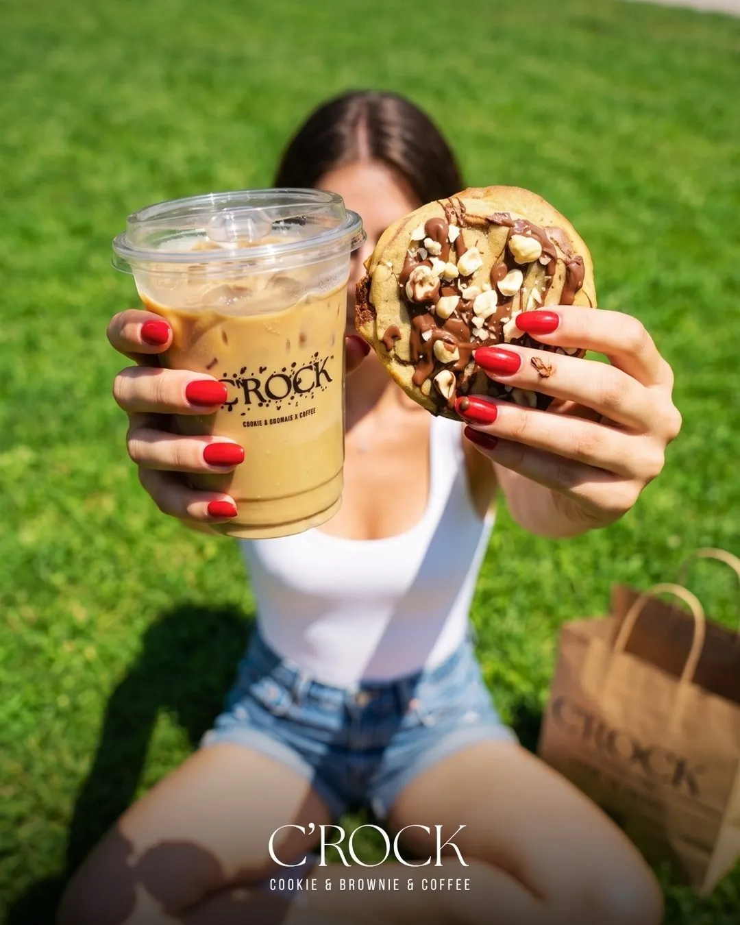 A woman on the grass holding an iced coffee in one hand and a C'ROCK cookie in the other