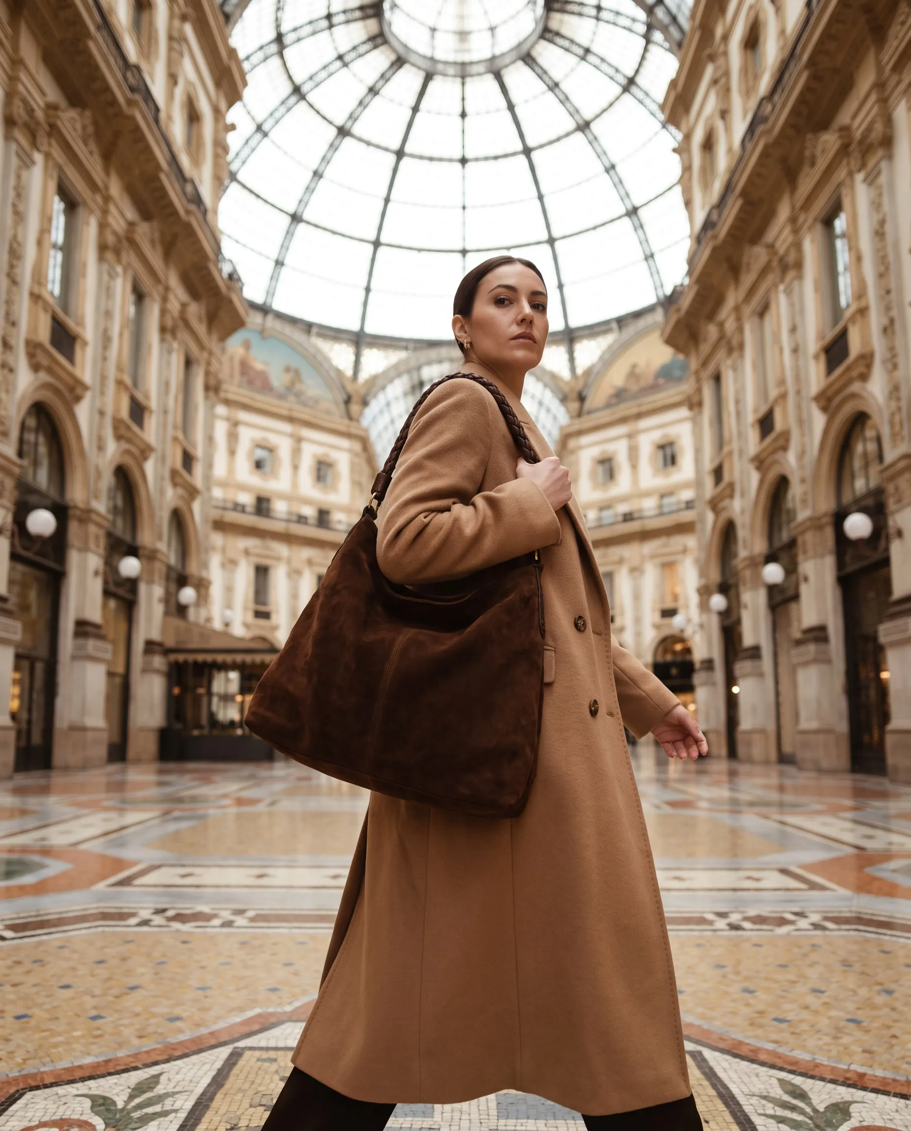 A model in a camel coat with a suede bag in a glass-roofed arcade